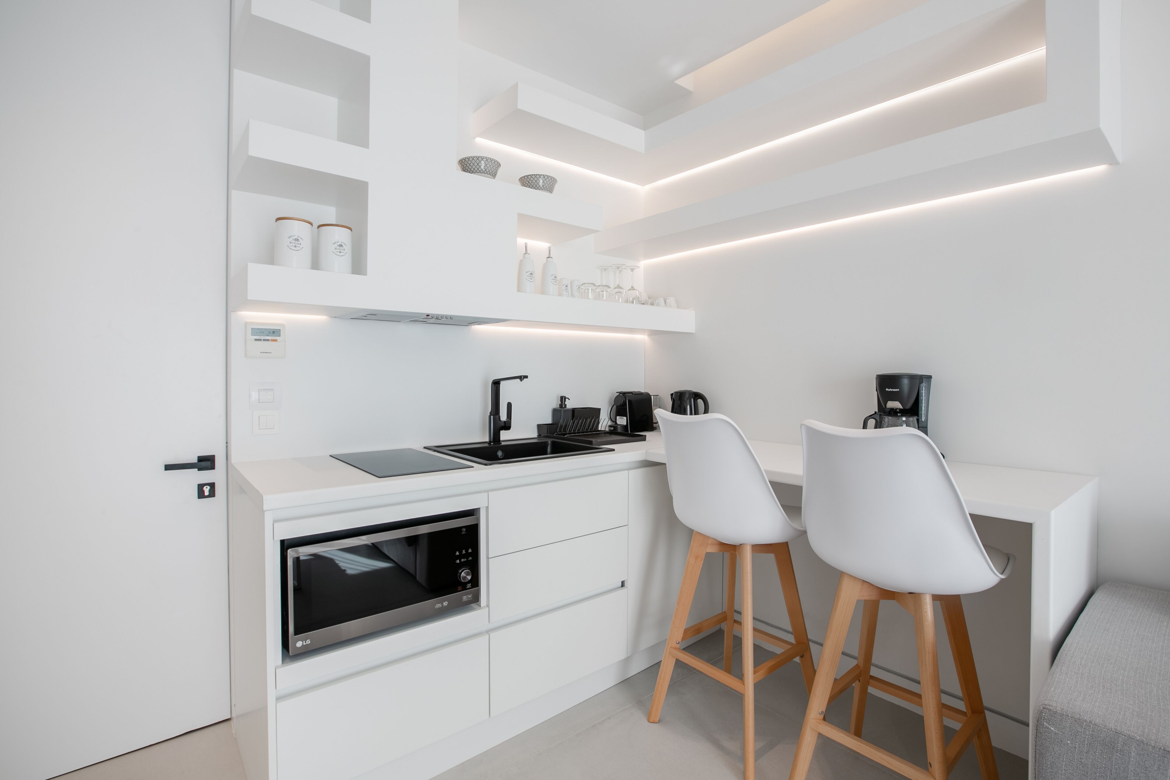 A modern kitchen with white cabinetry, a built-in oven, a sink, and a coffee machine. There are two white bar stools and a white countertop.