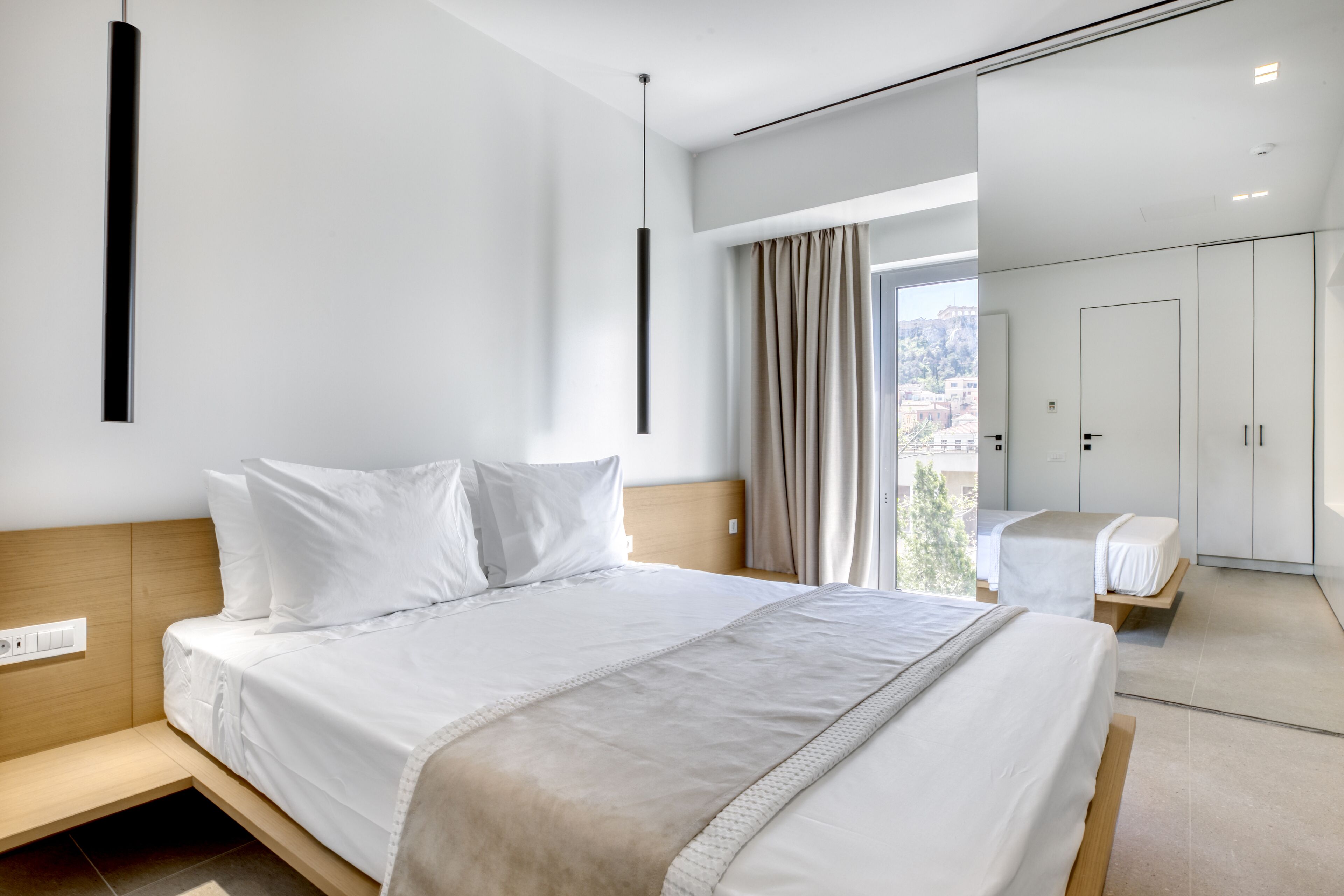 A modern hotel room with a large bed, a bedside table, a wardrobe, and a view of the city.