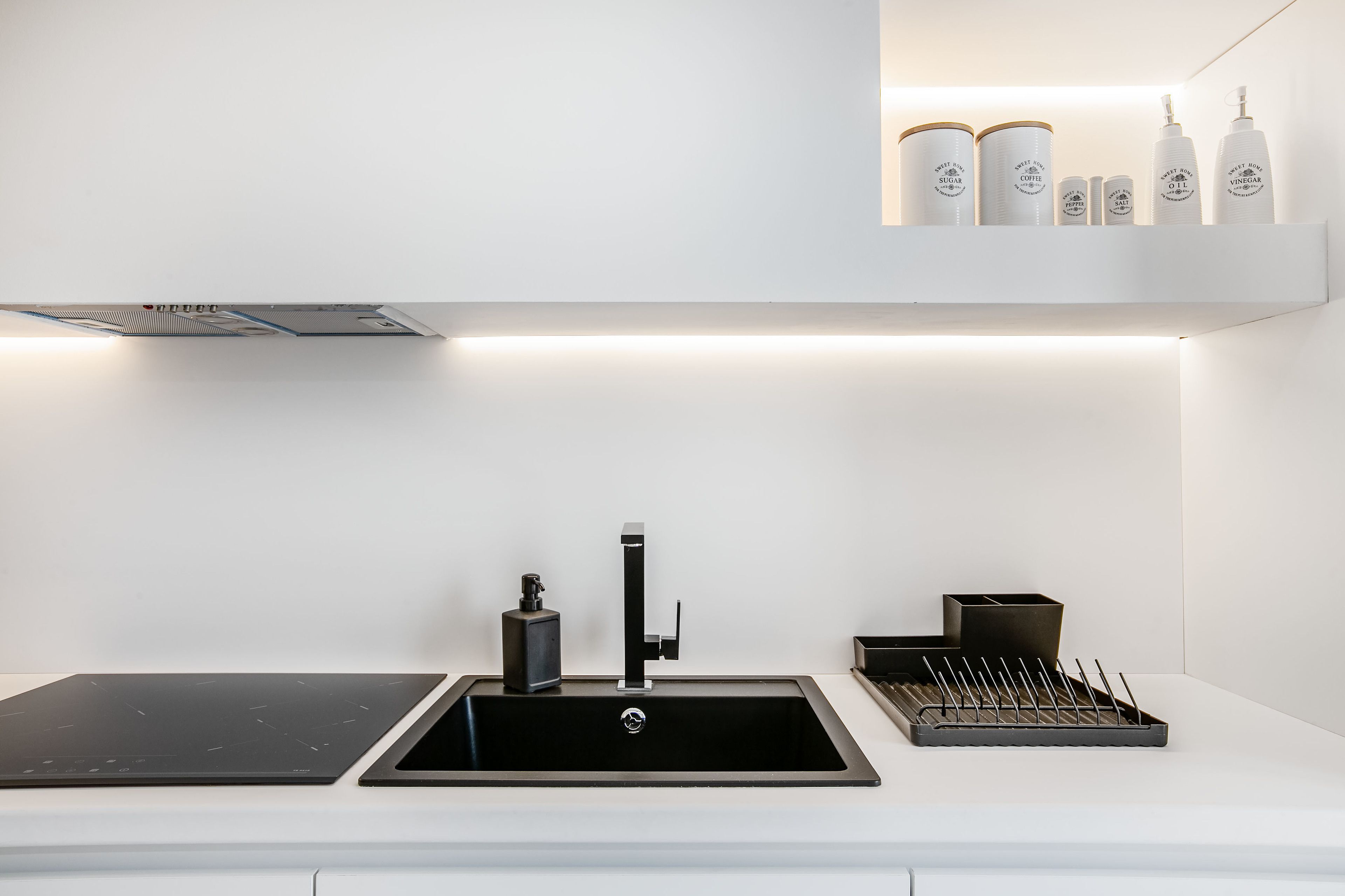 A modern kitchen with a black sink, a built-in stove, and a shelf above the sink holding various bottles.