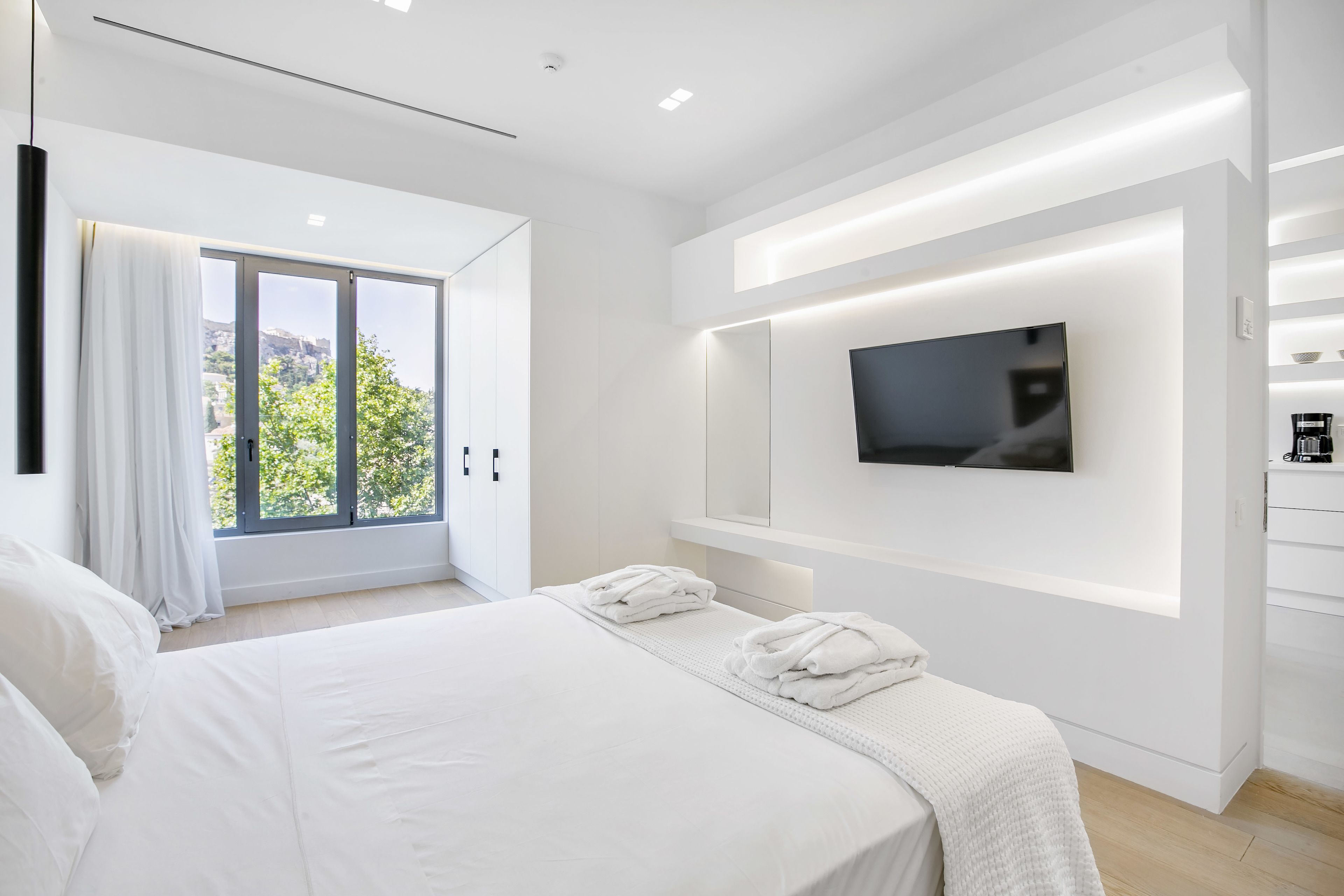 A modern hotel room with a large bed, a TV mounted on the wall, and a view of greenery outside.