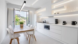 Una cocina moderna con armarios blancos, una mesa de comedor de madera y sillas. Se observa vegetación y edificios a través de la ventana.