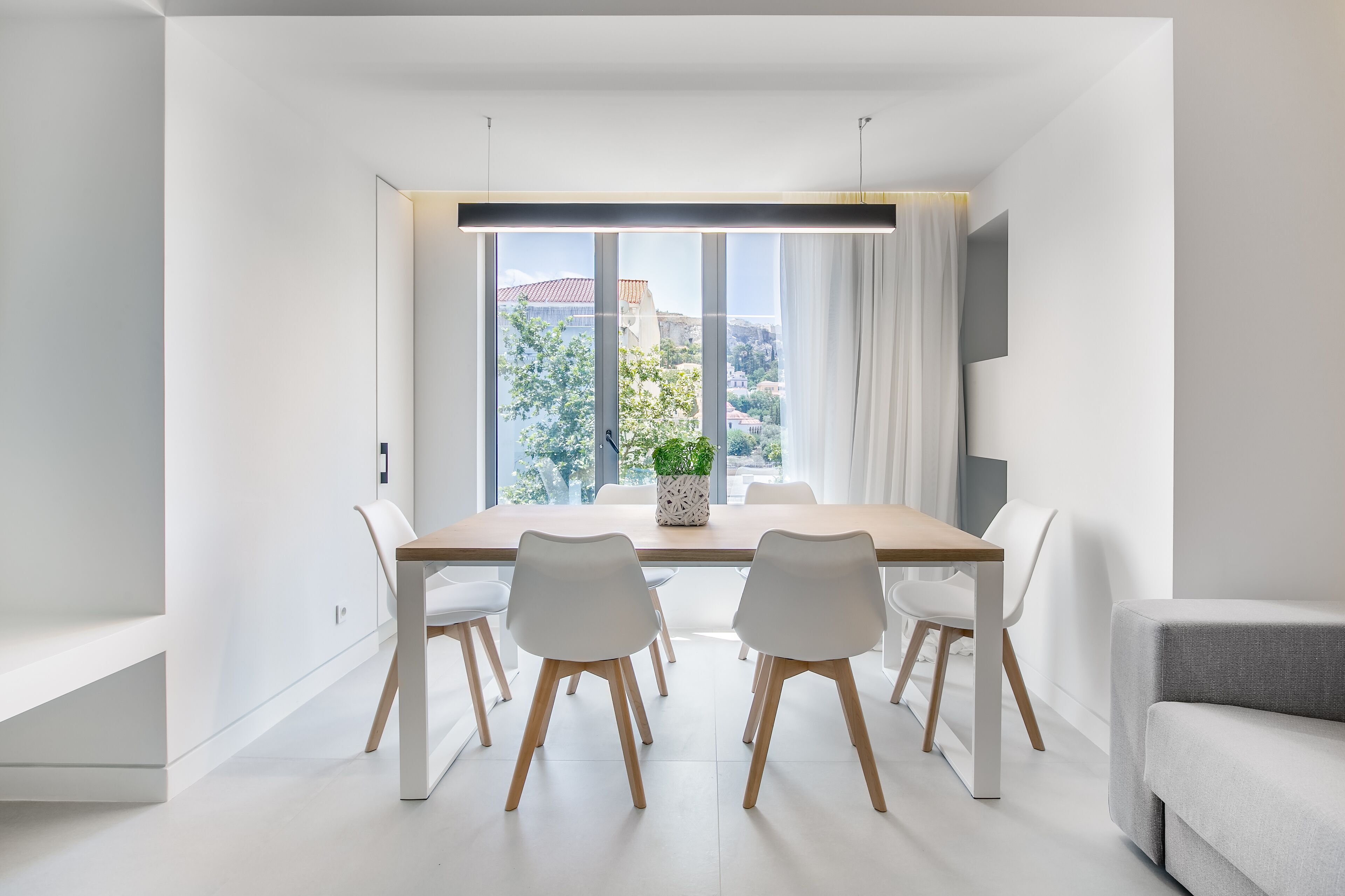 A modern dining area with a wooden table, white chairs, and a view of the city through large windows.