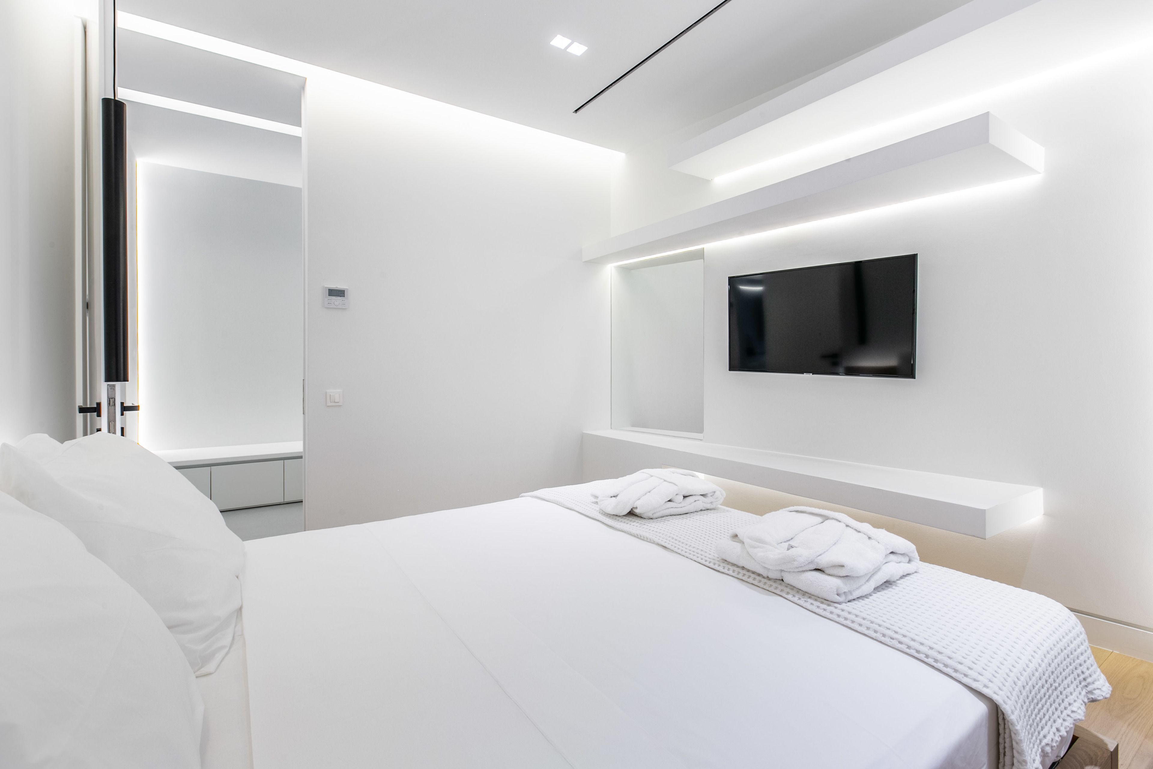 A modern hotel room with a large bed, a flat-screen TV mounted on the wall, and a built-in bench under the TV.