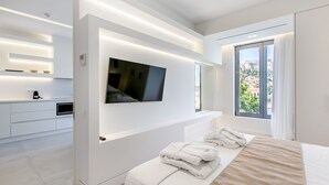 Una habitación de hotel moderna con una cama grande, un televisor de pantalla plana montado en la pared, una pequeña cocina y un ventanal con vistas a la ciudad.