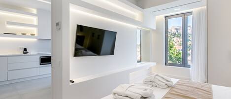 A modern hotel room with a large bed, a flat-screen TV mounted on the wall, a small kitchen area, and a large window offering a city view.