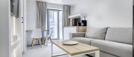 Uma sala de estar moderna e bem iluminada, com um sofá, uma mesa de centro de madeira e uma mesa de jantar com cadeiras. É visível uma área de cozinha ao fundo, com uma cafeteira e armários.