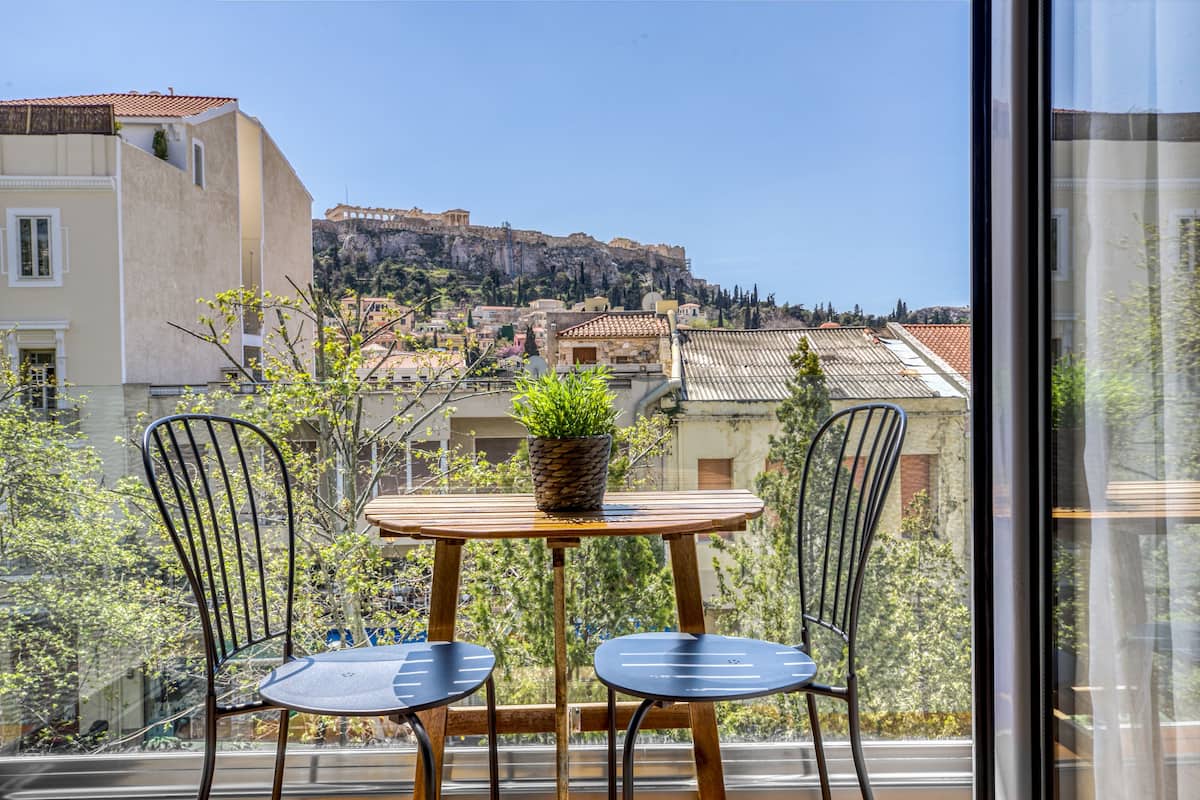Un balcon avec une table en bois et deux chaises, d’où l’on peut admirer un paysage urbain avec des bâtiments historiques et une falaise.