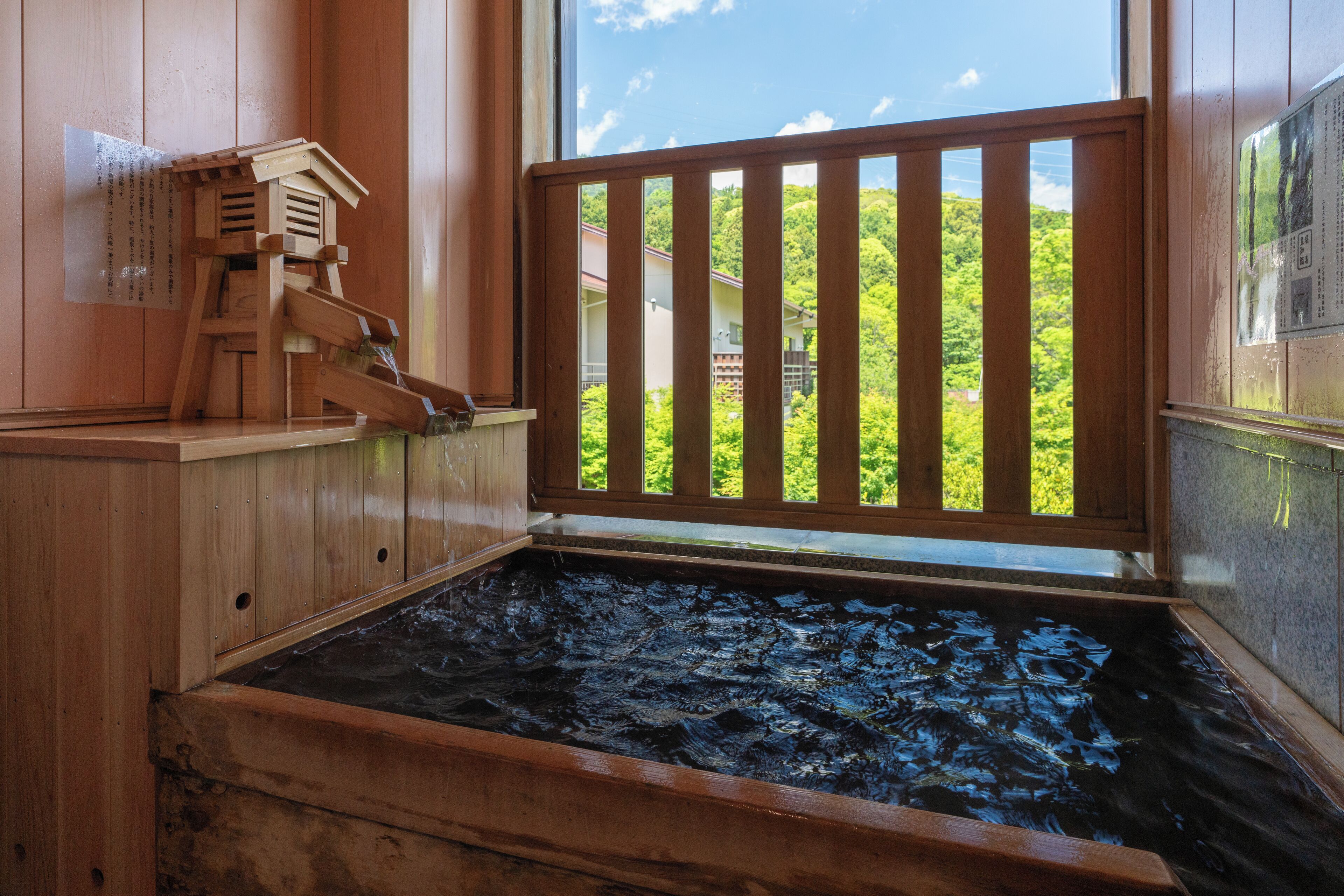 A wooden hot spring bath with a wooden railing and a view of a green landscape through a window.