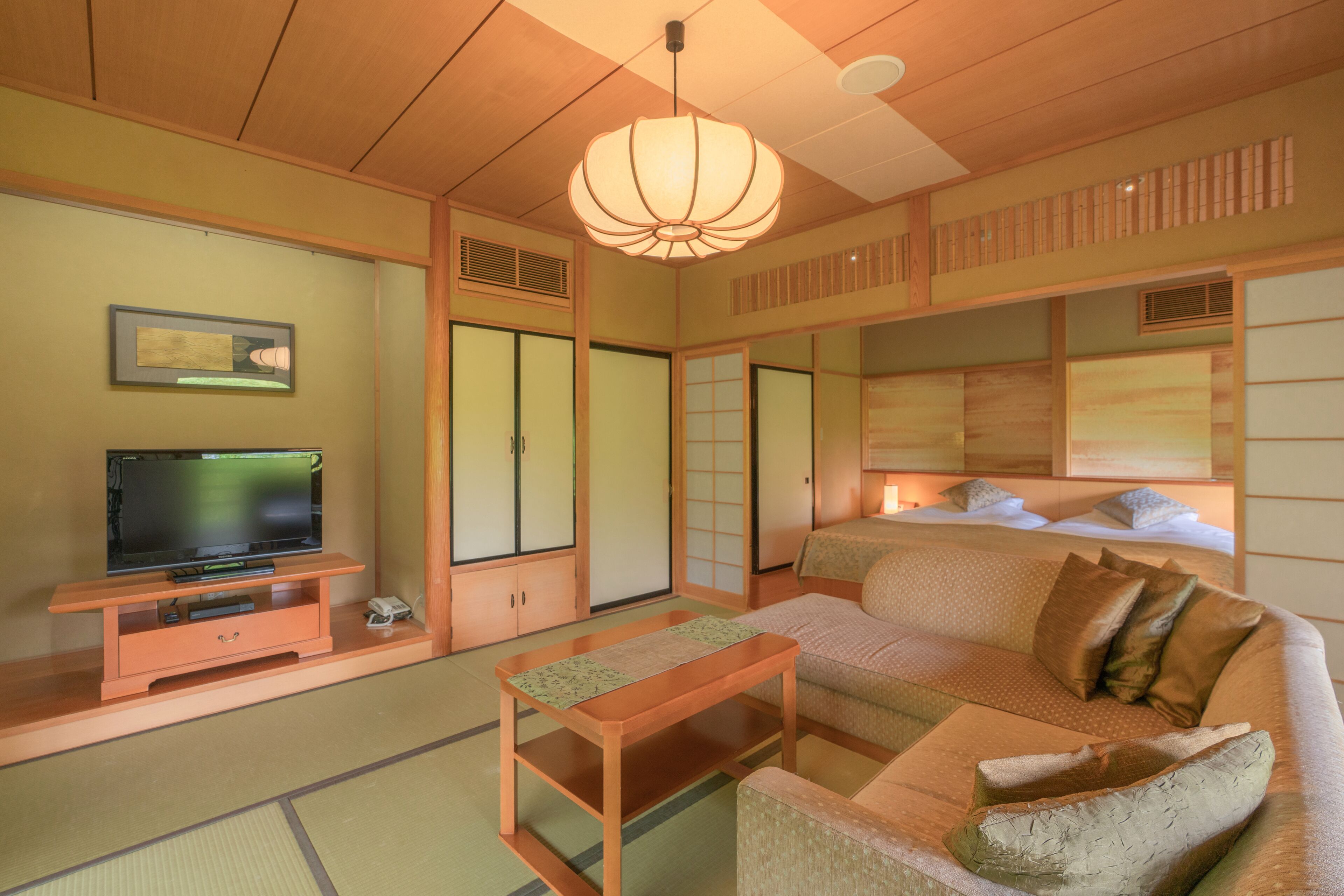A traditional Japanese-style living room with tatami flooring, a low wooden table, a sofa with cushions, a television on a wooden stand, sliding shoji doors, and a hanging paper lantern.