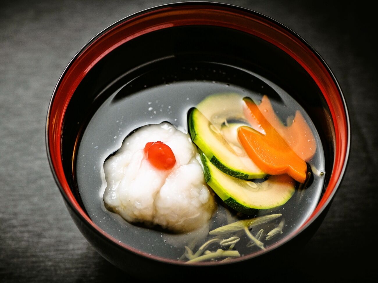 Uma tigela de sopa transparente com um bolinho branco, pepino fatiado, cenoura e um enfeite vermelho.