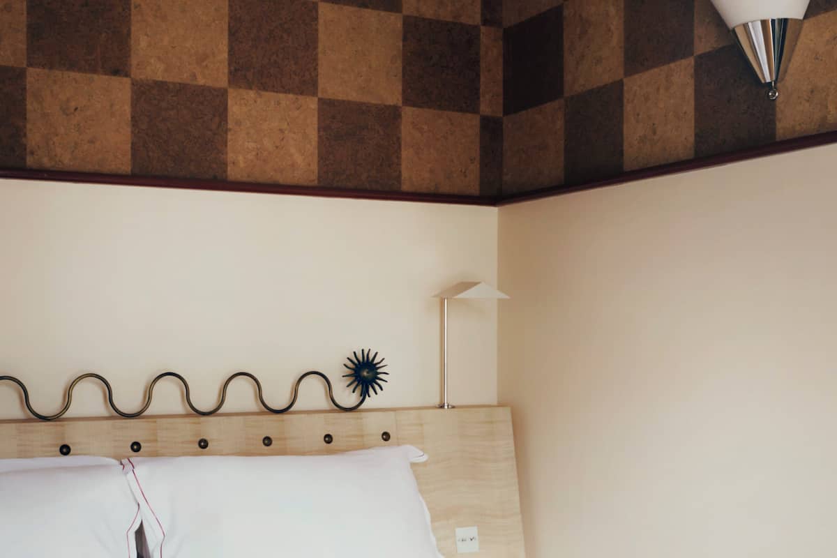 A hotel room with a checkered ceiling, a bed with white linens, a wall-mounted lamp, and a wooden headboard.