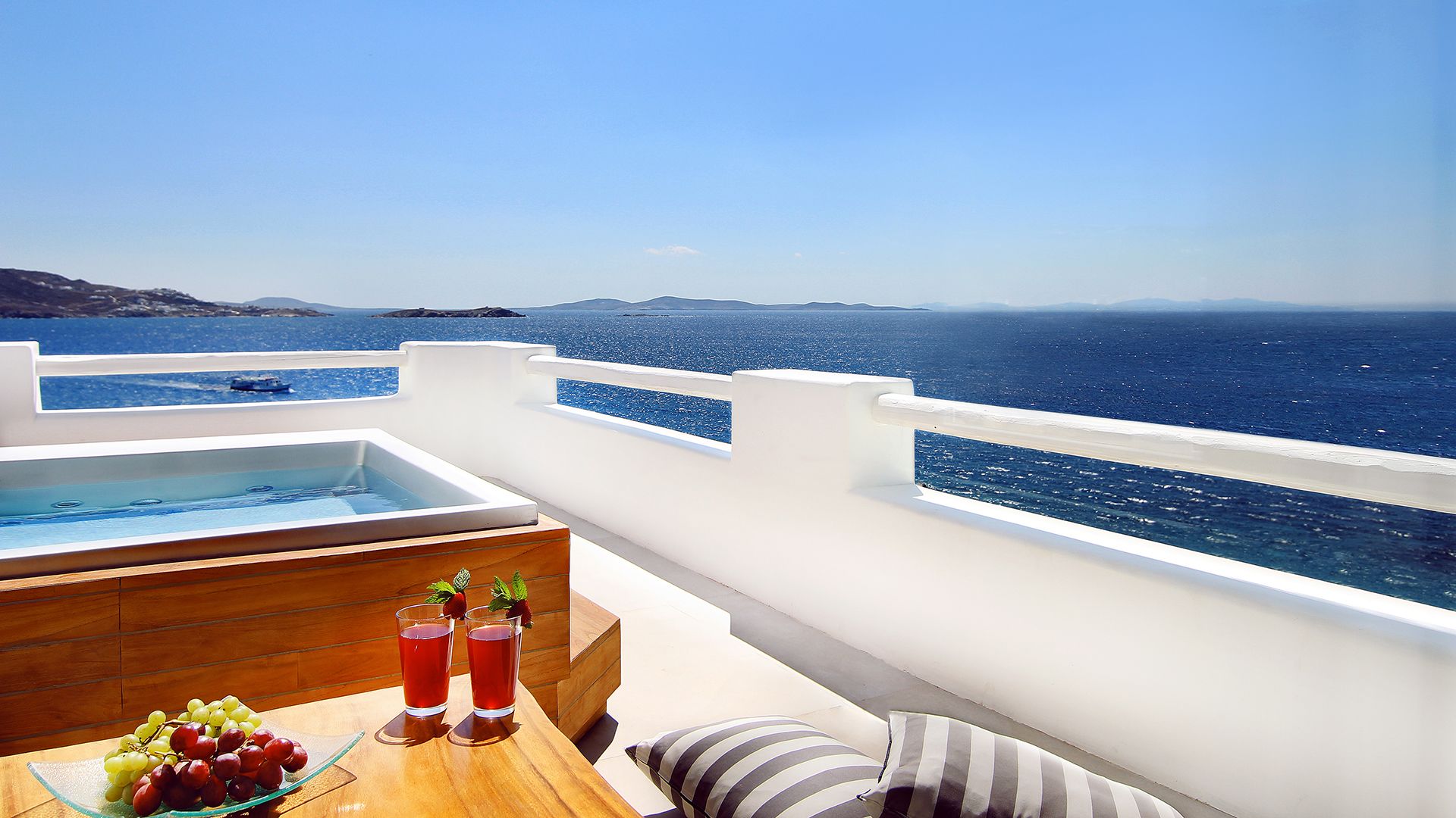 Een balkon met een jacuzzi, een houten tafel met een fruitschaal en drankjes, en uitzicht op de zee en verre eilanden.