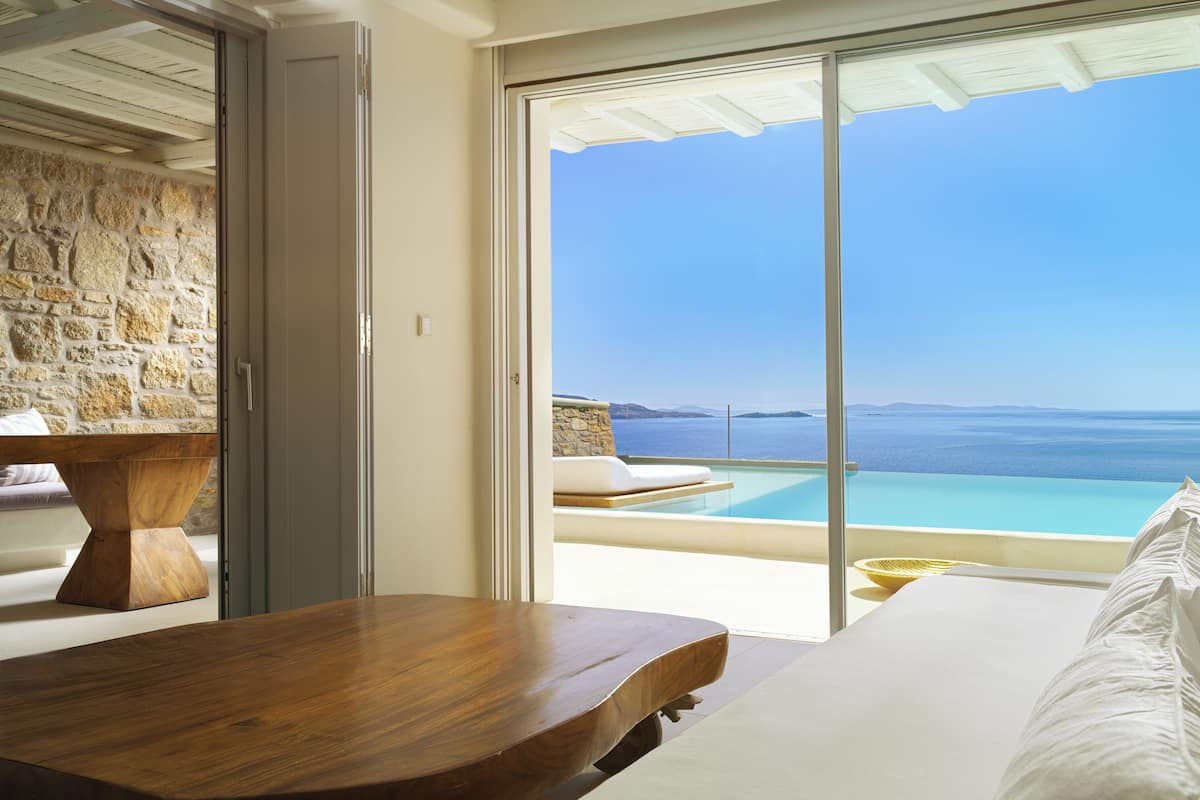 A room with a wooden table, a bed, and a view of the sea through large windows.