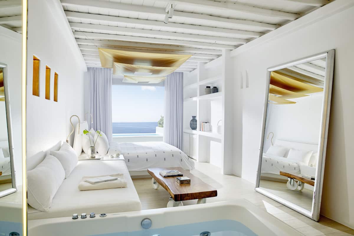 A modern bedroom with a large bed, a small sofa, a wooden side table, and a large mirror.
