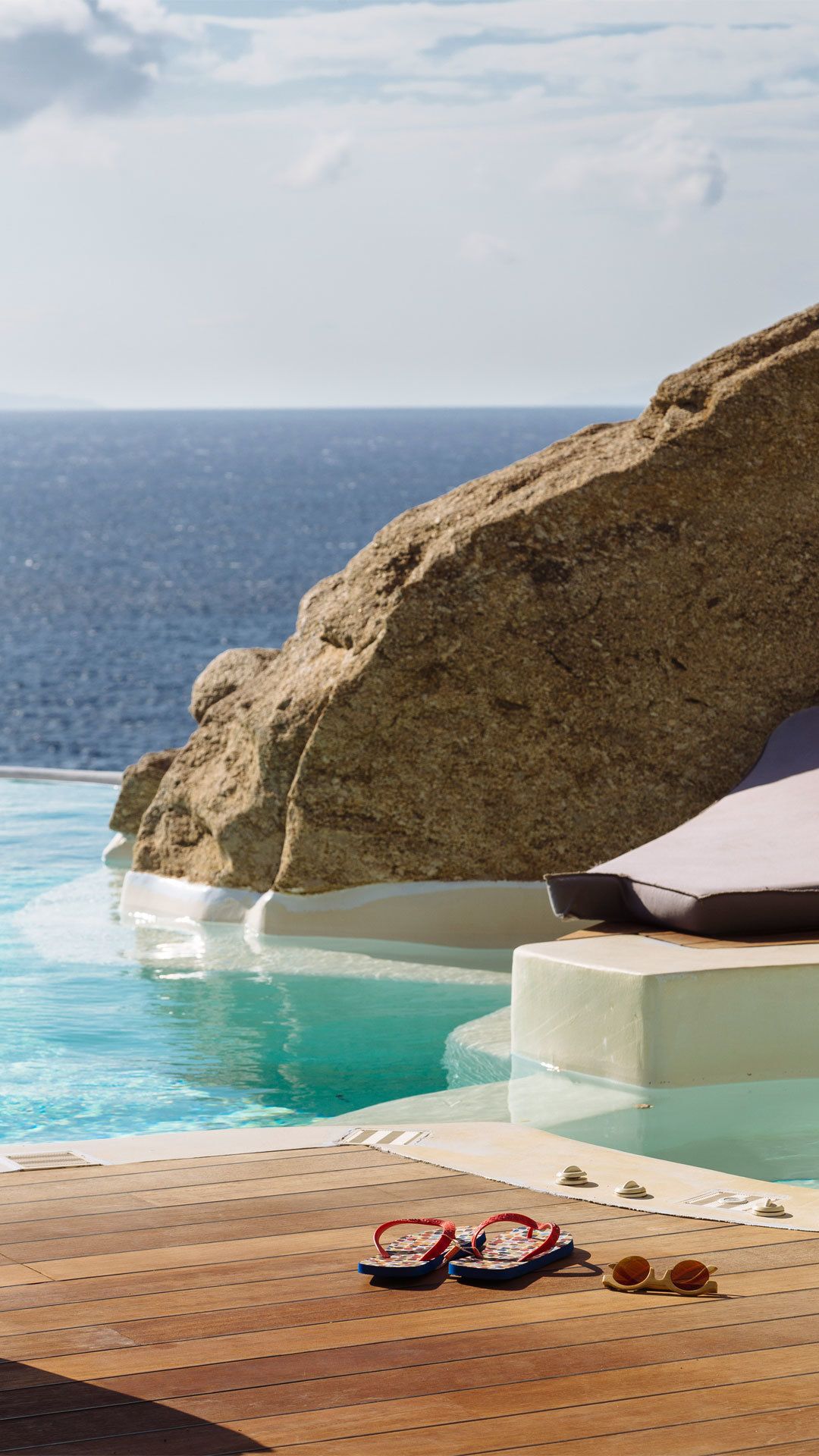 Una piscina con agua azul cristalina, una gran formación rocosa y una terraza de madera con chanclas y gafas de sol.