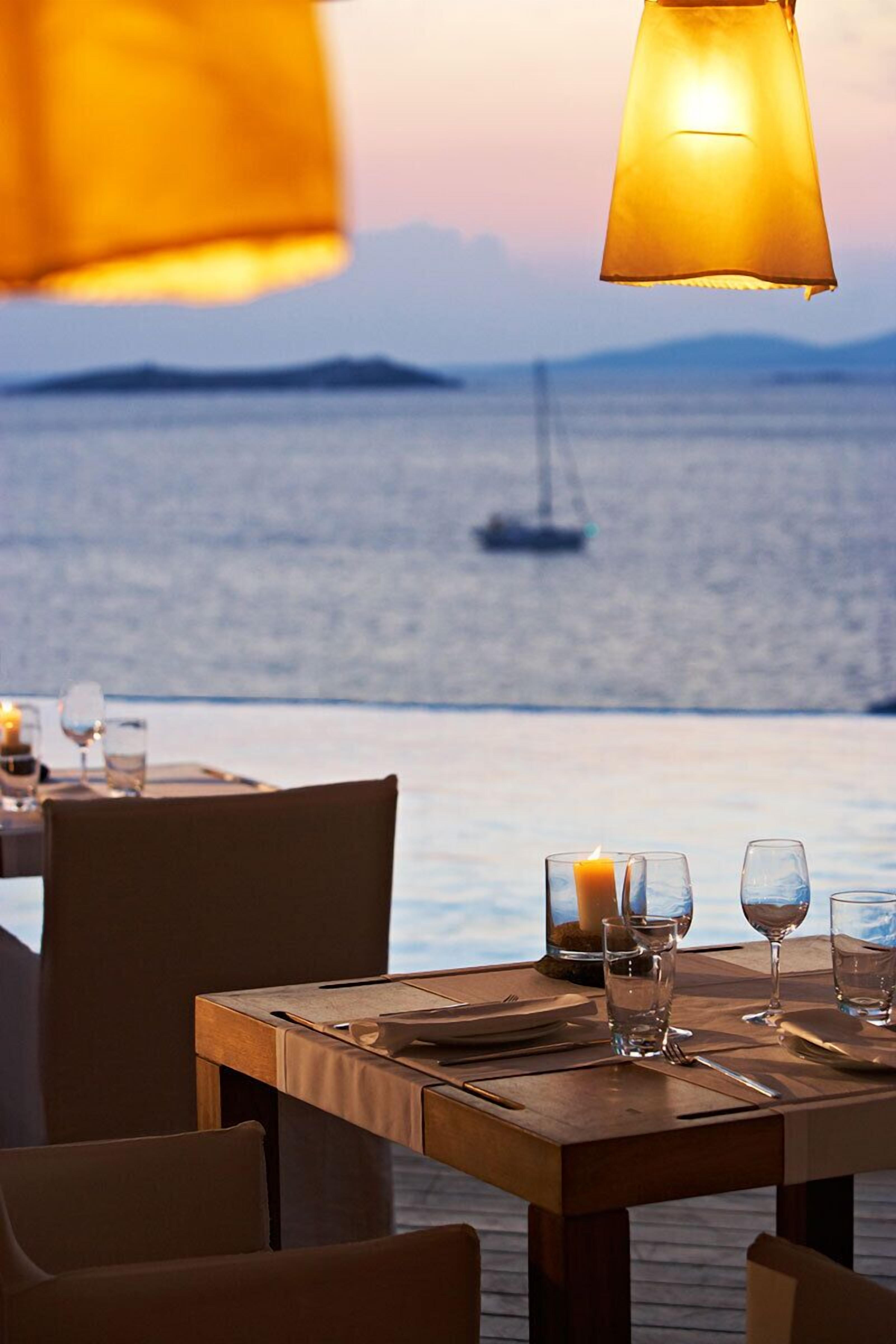 Mesa para dos dispuesta con vela, vasos y cubiertos, con vista a un mar tranquilo y un velero a lo lejos.