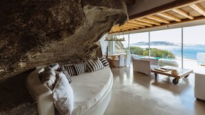 A modern living room with a large rock feature, a white sofa, and a view of the sea. - CAVO TAGOO MYKONOS (Mykonos)