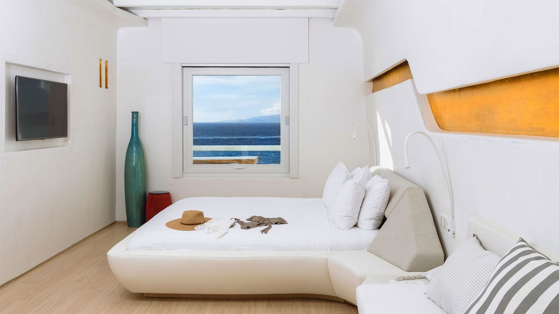 Un dormitorio moderno con un gran ventanal que ofrece vista al mar, una cama con ropa blanca y una pequeña mesa con un sombrero y una bufanda.