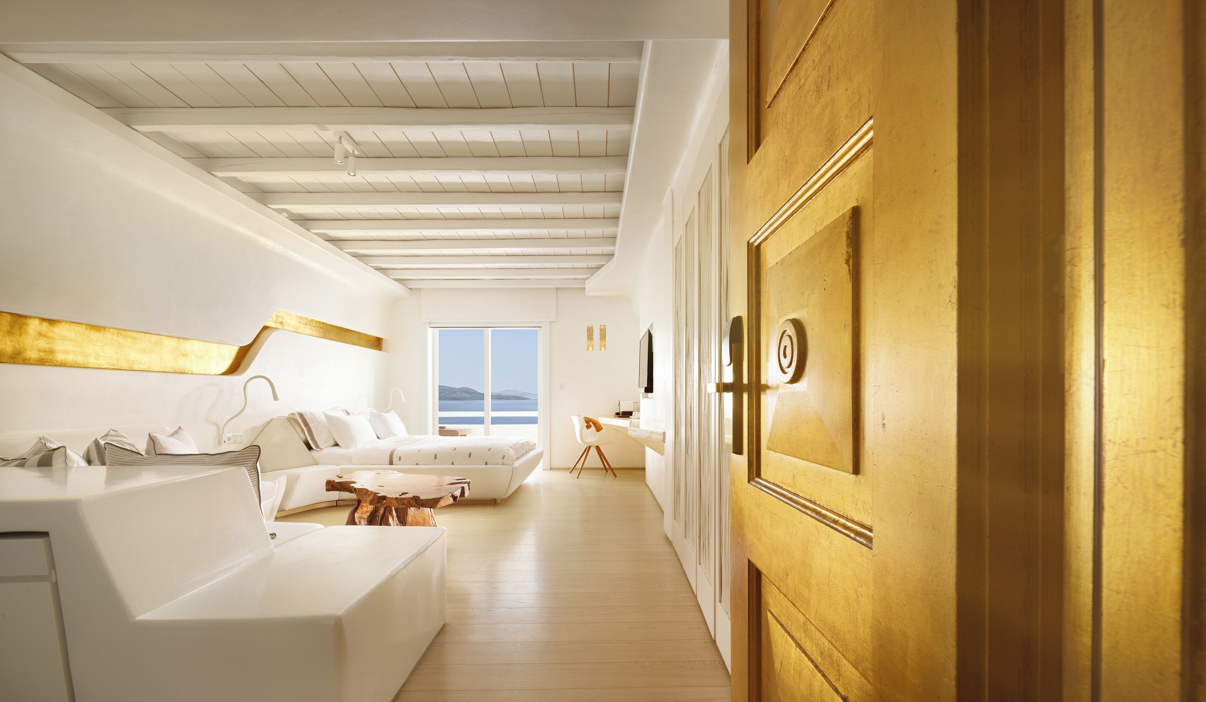 A modern hotel room with a wooden ceiling, a large window with a view of the sea, a white sofa, and a wooden door.