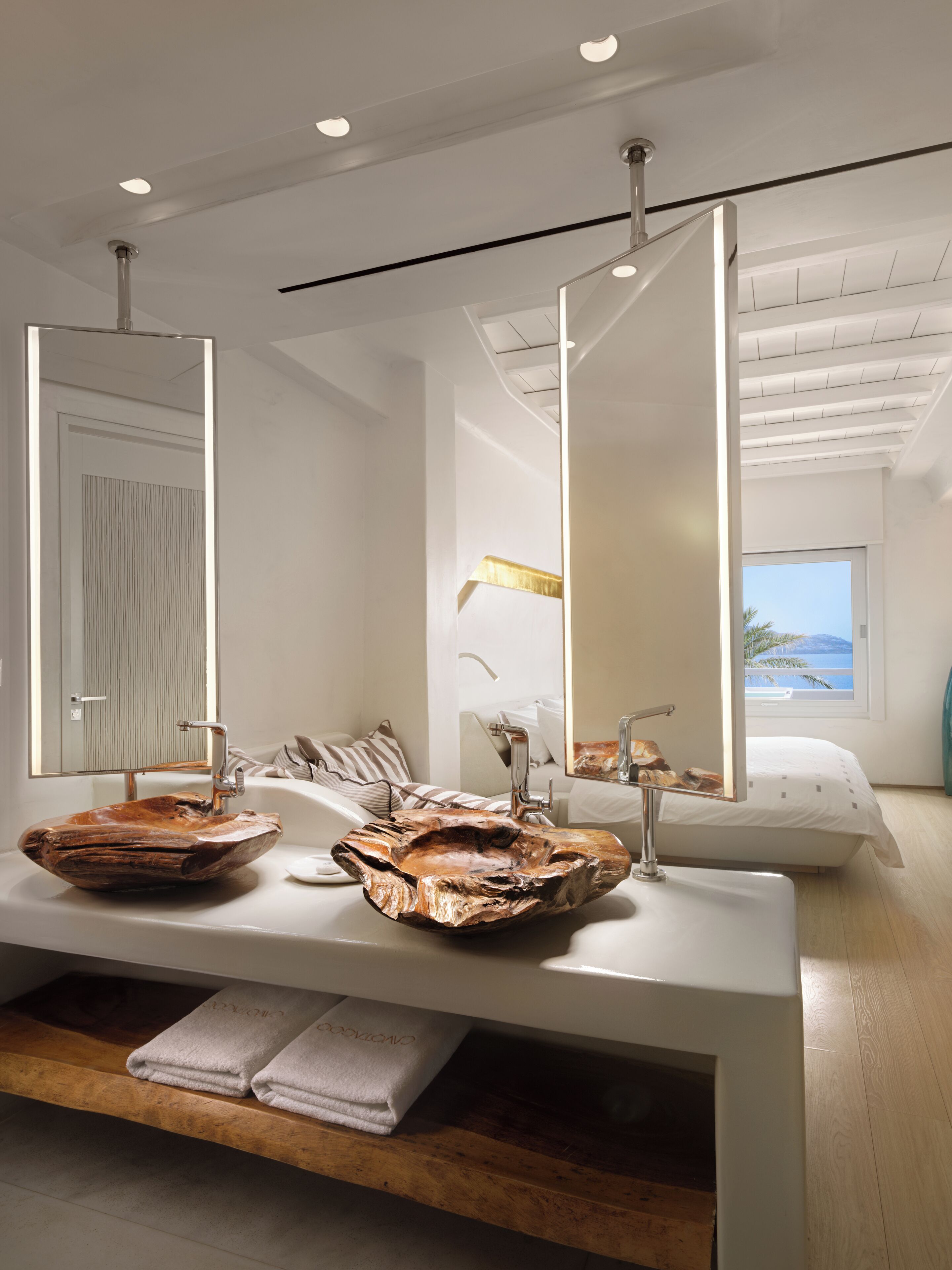 Un baño moderno con un espejo grande, dos lavamanos con cuencos de madera únicos y un banco de madera con toallas.