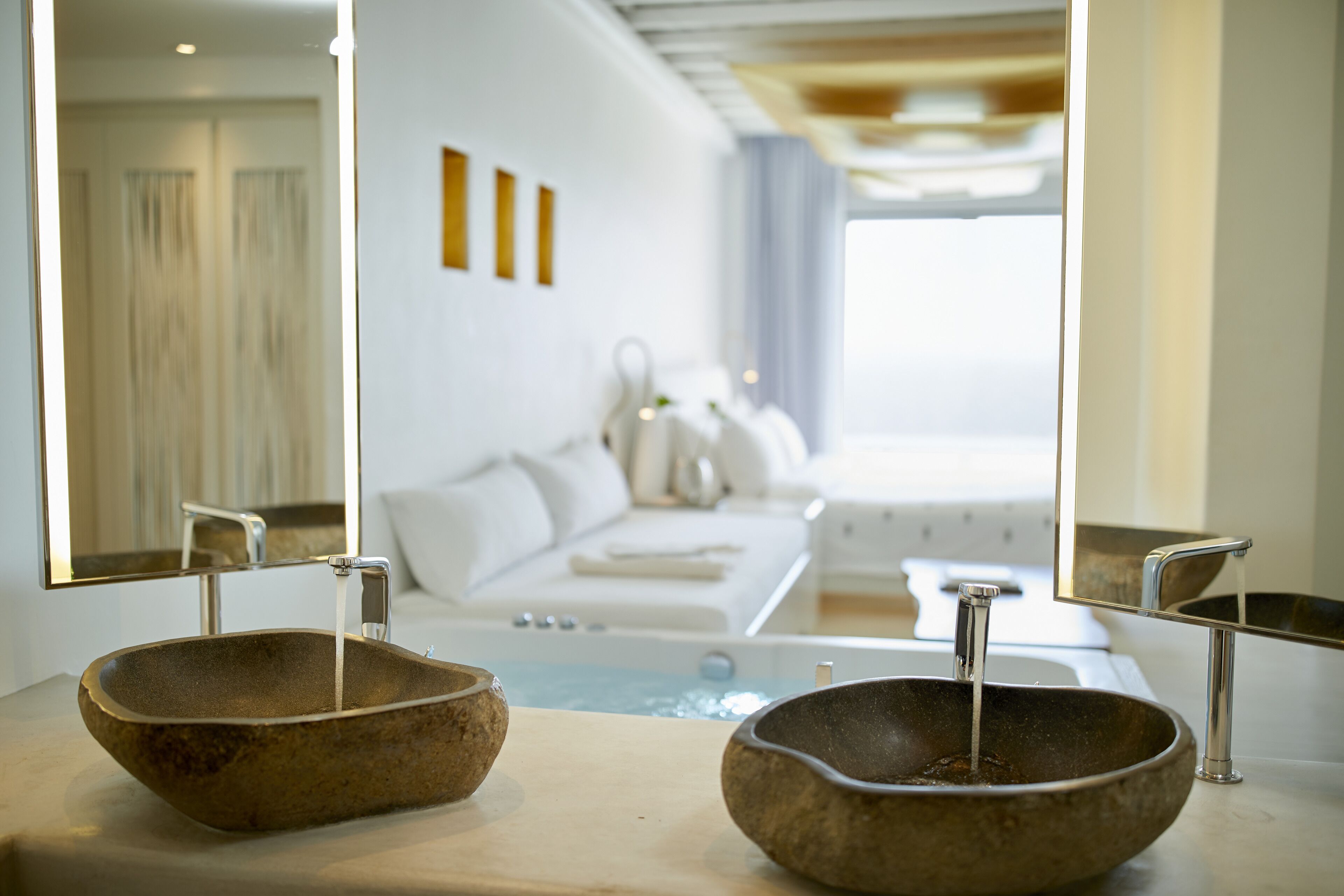 Baño con dos lavamanos de piedra y un espejo grande que refleja un dormitorio con cama y ventana.