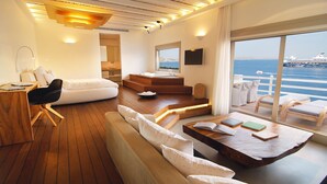 A modern interior with a wooden floor, white furniture, a large window with a view of the sea, and a balcony with white chairs. - CAVO TAGOO MYKONOS (Mykonos)