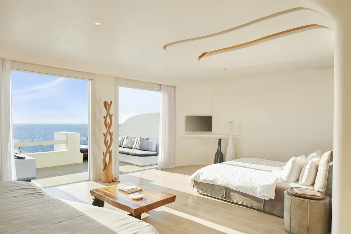 A modern bedroom with a large bed, a sofa, a wooden coffee table, and a view of the sea.