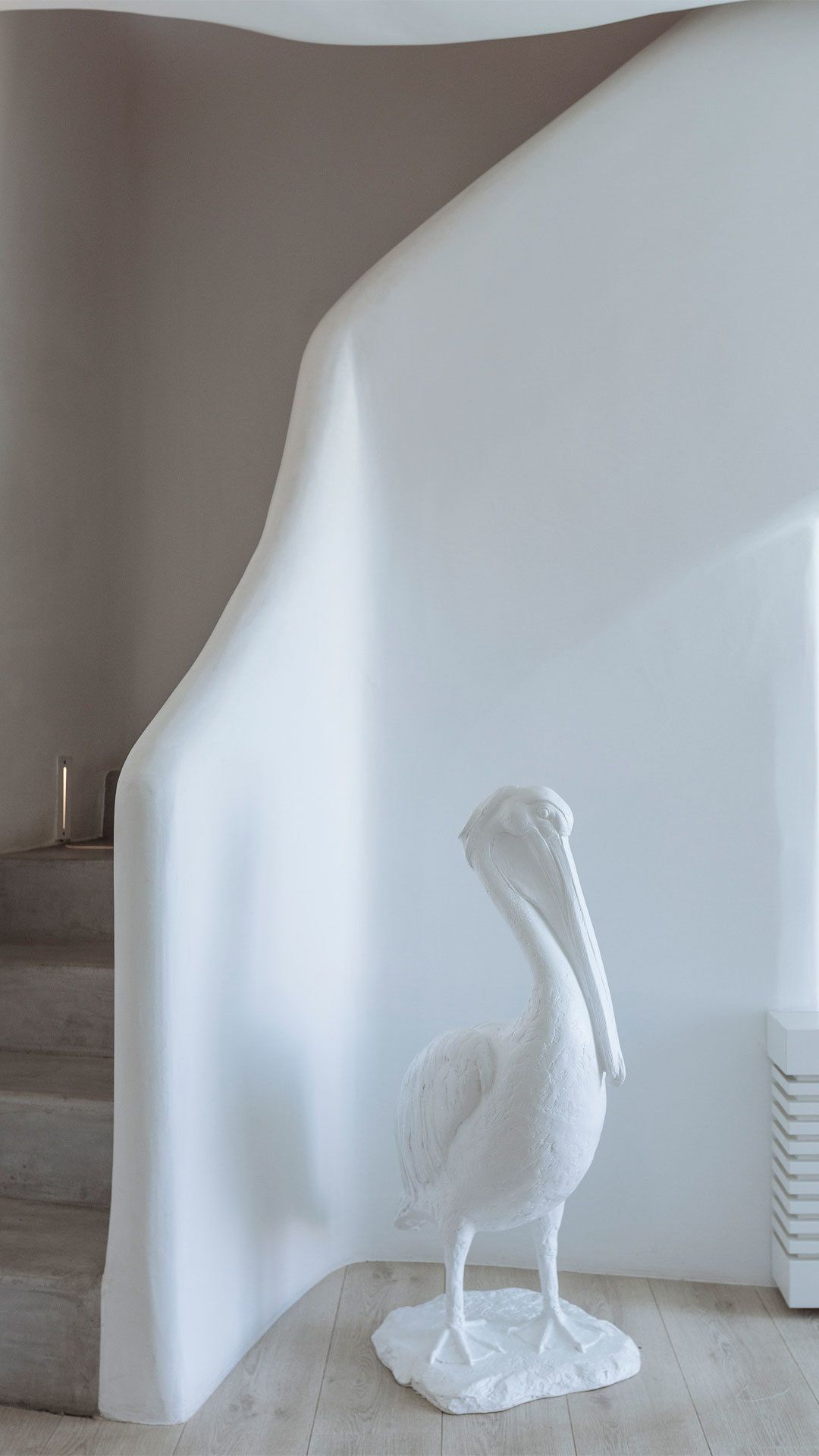A white sculpture of a pelican on a textured base, positioned in a corner of a room with a curved white wall and a staircase in the background.