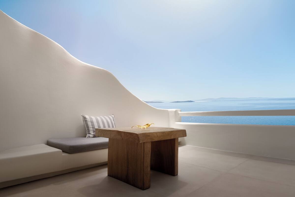 A modern room with a curved white wall, a wooden table, and a gray sofa with a striped pillow. A view of the sea and distant mountains is visible through a large window.