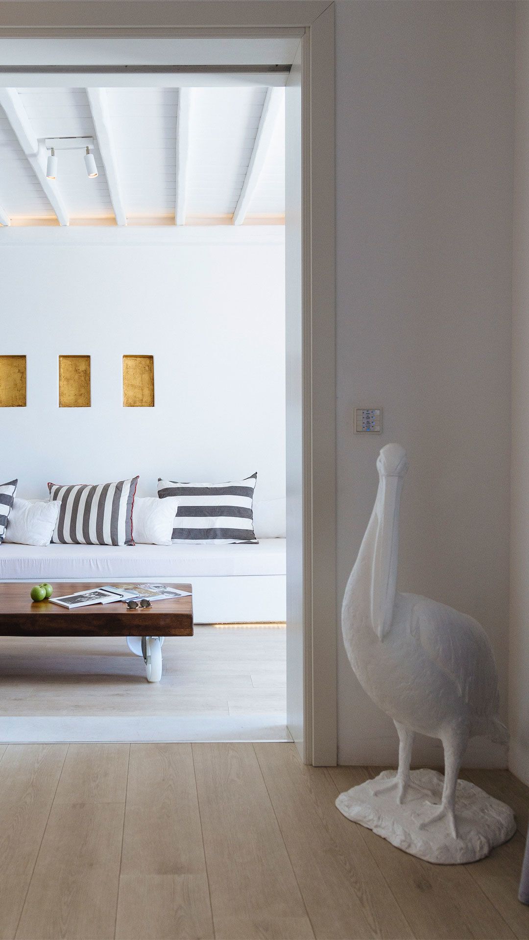 Een moderne woonkamer met een witte bank, gestreepte kussens, een houten salontafel met boeken en appels, en een witte pelikaan sculptuur.