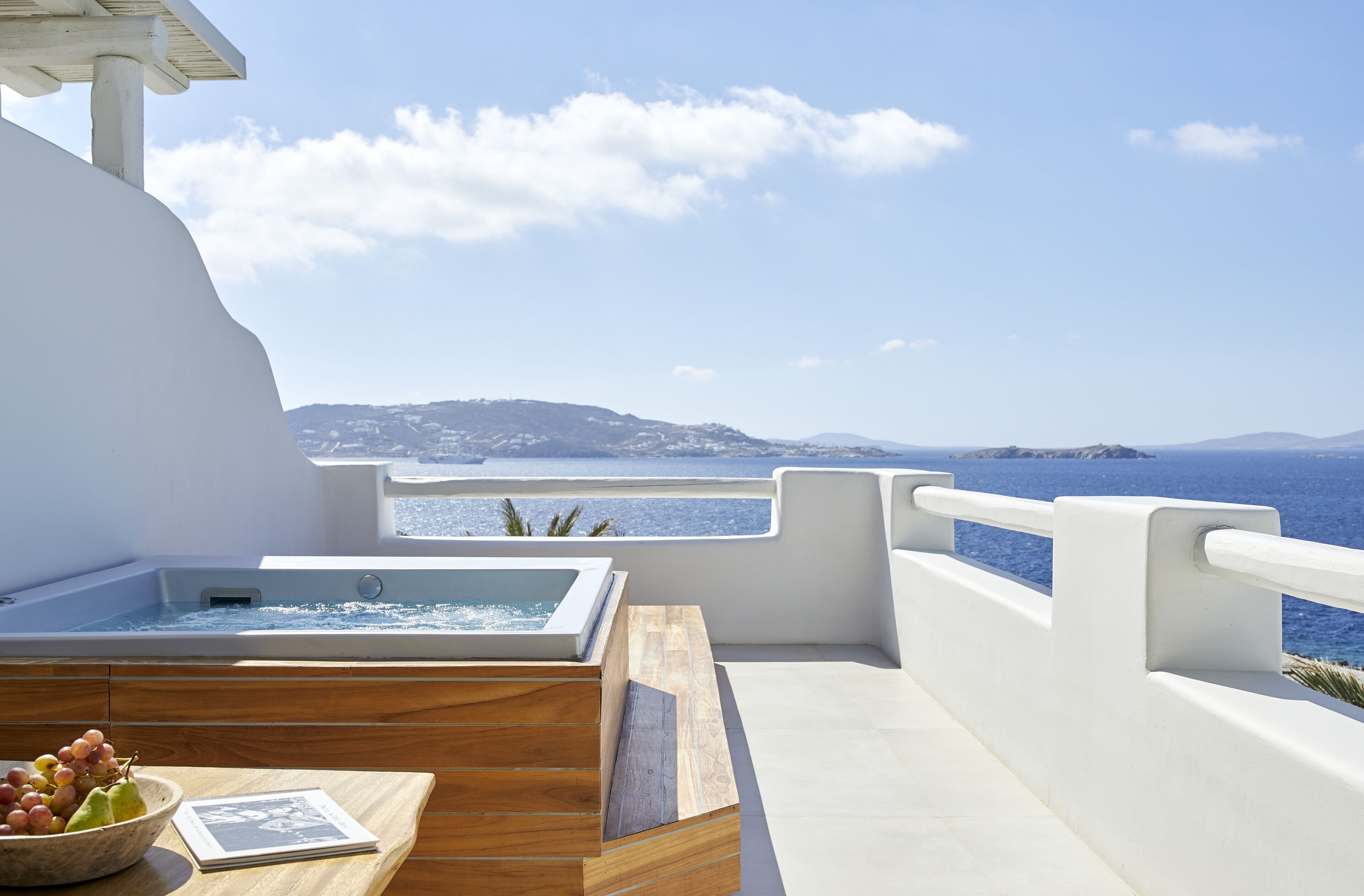 A modern outdoor seating area with a wooden table, a bowl of fruit, and a hot tub overlooking a scenic coastal view.