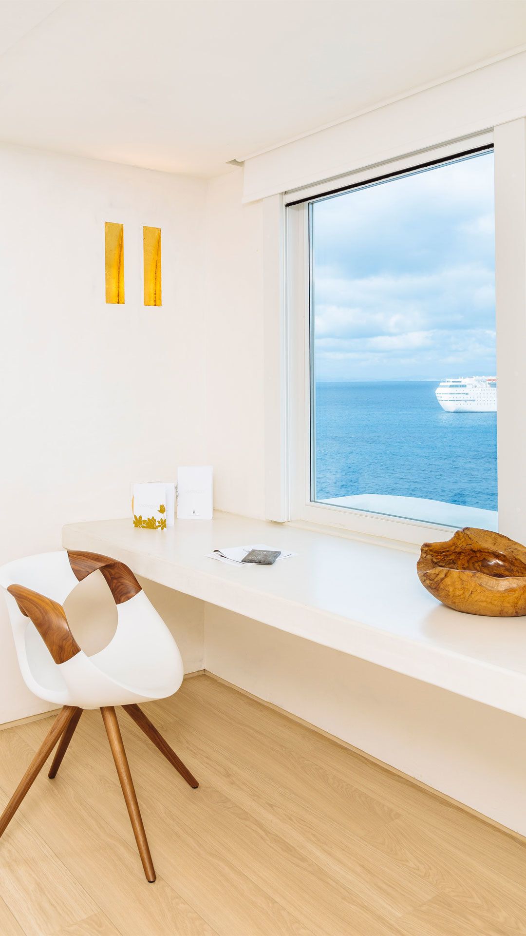 A modern room with a large window overlooking the sea, a white bench, a wooden bowl, and a white chair with wooden accents.