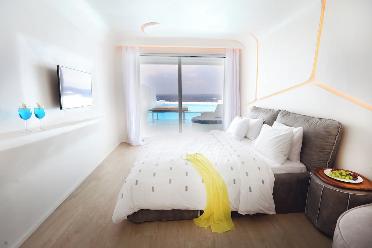 Une chambre d’hôtel moderne avec un lit, une petite table sur laquelle sont disposés des fruits, une télévision et une vue sur l’océan.