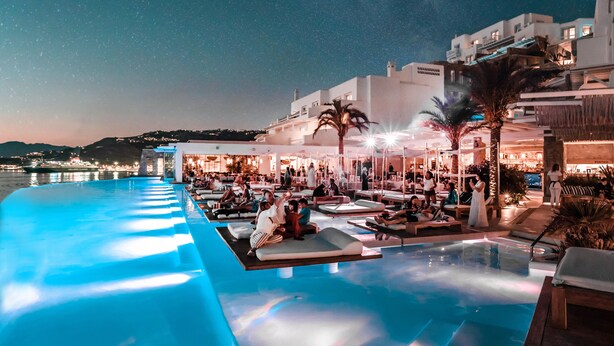 A poolside area with lounge chairs and a view of the sea at night. - CAVO TAGOO MYKONOS (Mykonos)