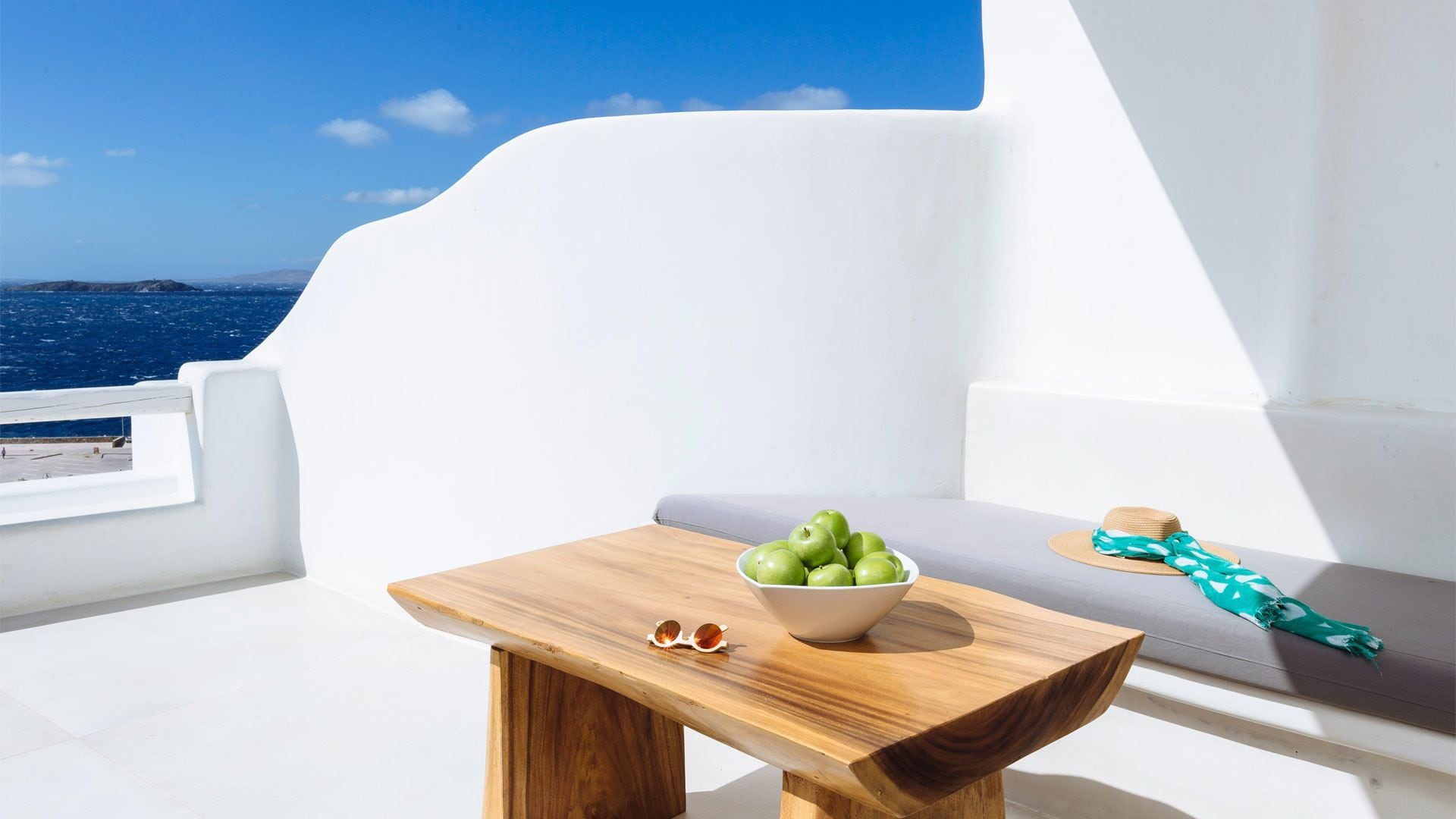 A wooden table with a bowl of green apples and a pair of sunglasses on a balcony with a white railing, overlooking the sea.