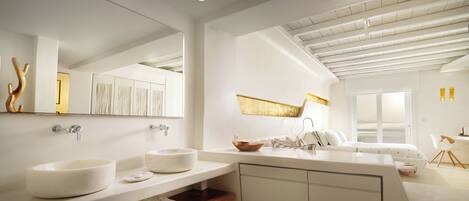 A modern bathroom with a large mirror, two sinks, a bathtub, and a shower. There is a wooden shelf with towels and a window providing a view of the outdoors.
