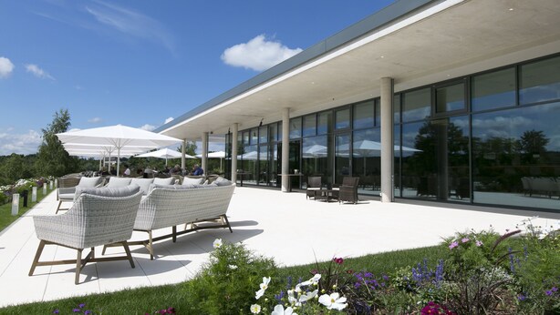 An outdoor seating area with wicker furniture and umbrellas, a modern building with large windows, and a garden with various flowers.
