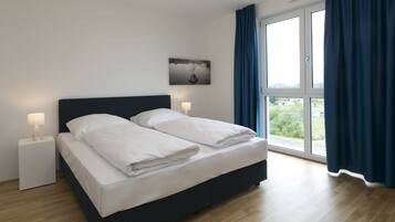 Una habitación de hotel moderna con una cama grande, dos mesitas de noche con lámparas, vistas a zonas verdes por la ventana y un cuadro en la pared.