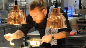 A person cooking in a kitchen with copper pendant lights, stainless steel appliances, and a checkered cloth.