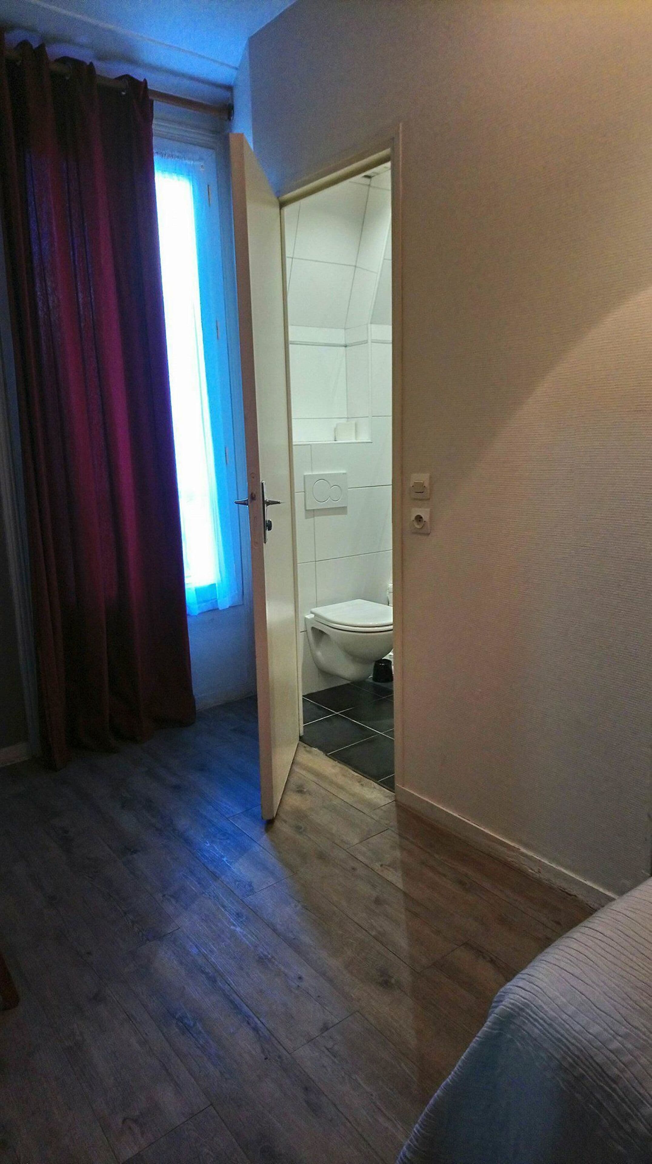 Een kamer met een houten vloer, een deur naar een badkamer met een wit toilet, en een raam met gordijnen.