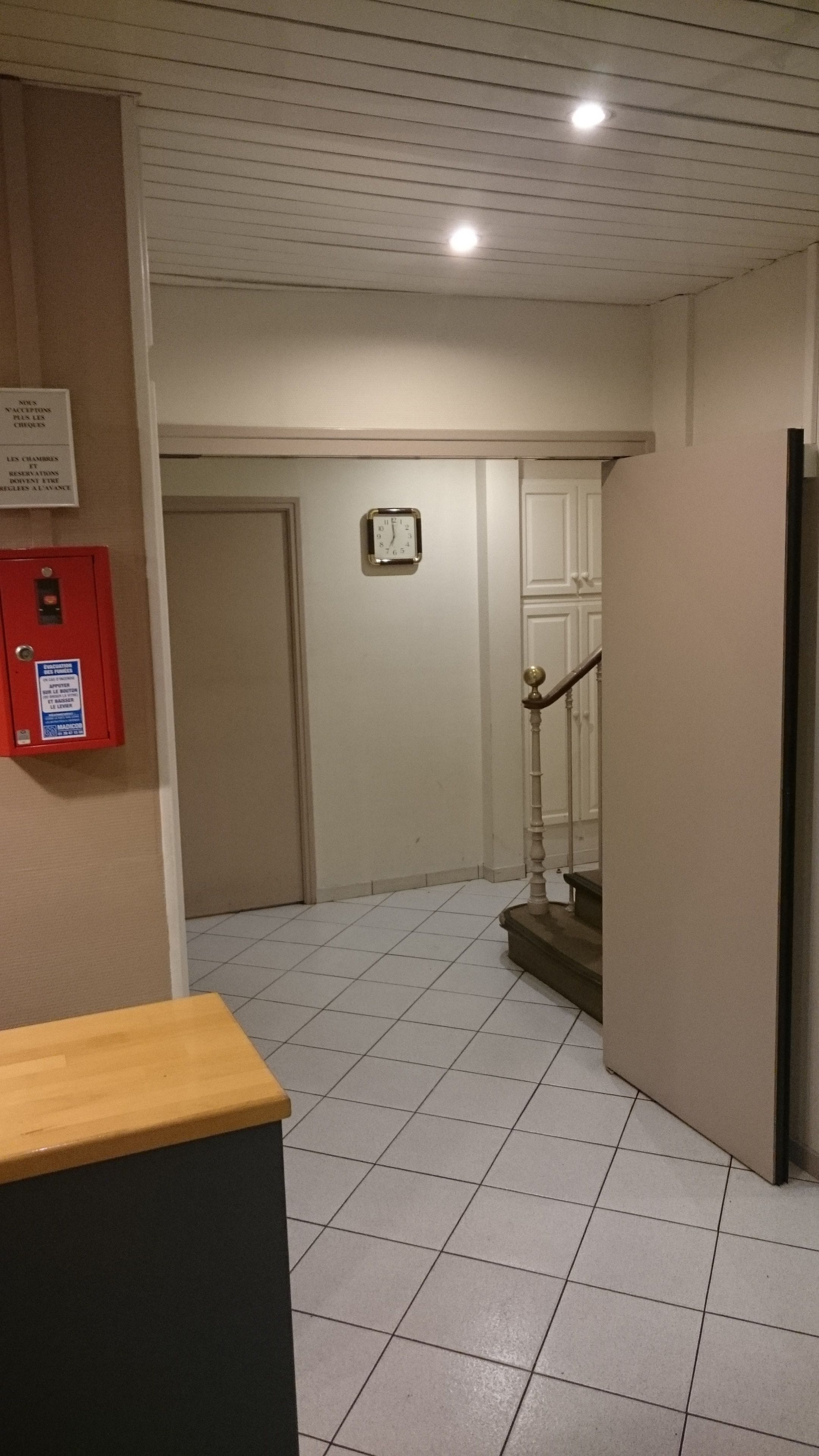 A hotel lobby with a tiled floor, a red fire alarm box, a clock on the wall, and a staircase leading to another floor.