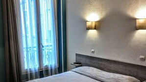 A hotel room with a large bed, a nightstand, two wall-mounted lights, and a window with curtains. - Acacia Hotel (Paris)