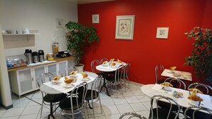 A dining area with tables set for breakfast, including croissants and coffee cups. There are chairs, a potted plant, and a red wall with framed pictures. - Acacia Hotel (Paris)