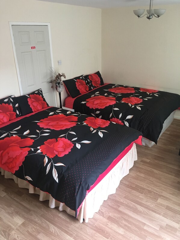 A bedroom with two beds, each with a black bedspread featuring red floral patterns and white accents. There is a white door with a red sign, a small plant, and a ceiling light fixture.