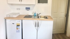 Une cuisine de taille réduite, avec des meubles blancs, un évier et un réfrigérateur. Un panneau d’affichage sur le mur rappelle de maintenir les lieux propres.