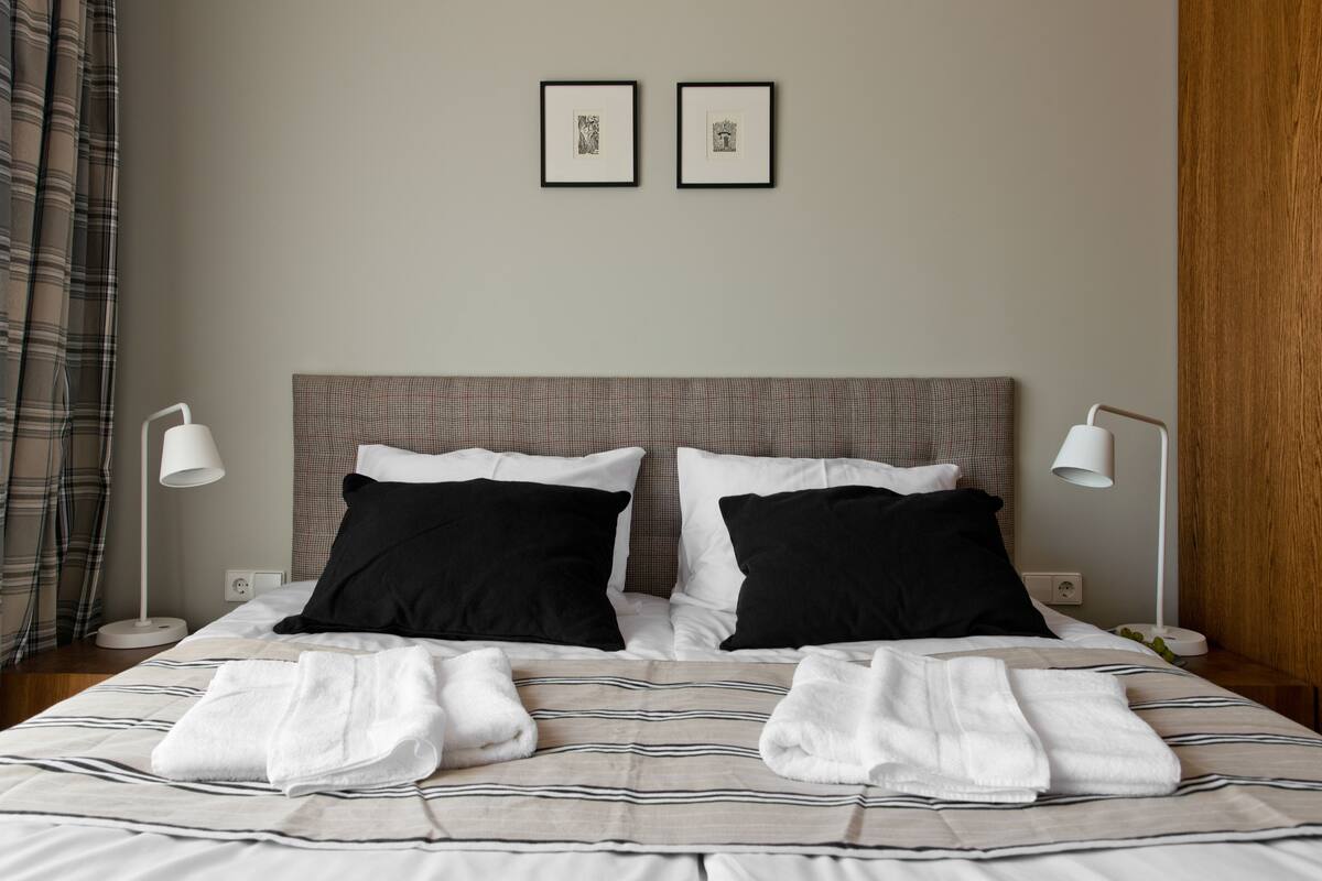 Een net opgemaakt bed met zwart-wit kussens, twee ingelijste foto’s aan de muur, en een bedlampje aan beide zijden.