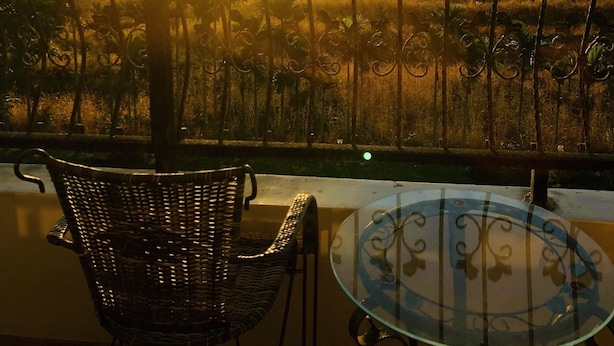 A balcony with a wicker chair and a glass table, overlooking a sunset over a rural landscape with fields and distant buildings. - Happiness House (Chenggong)