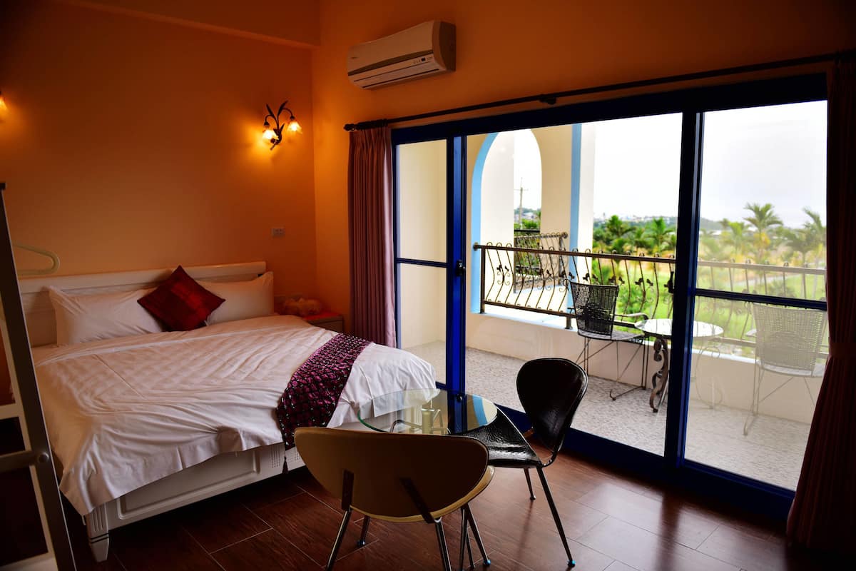 A hotel room with a bed, a glass table, a chair, and a balcony with outdoor furniture.