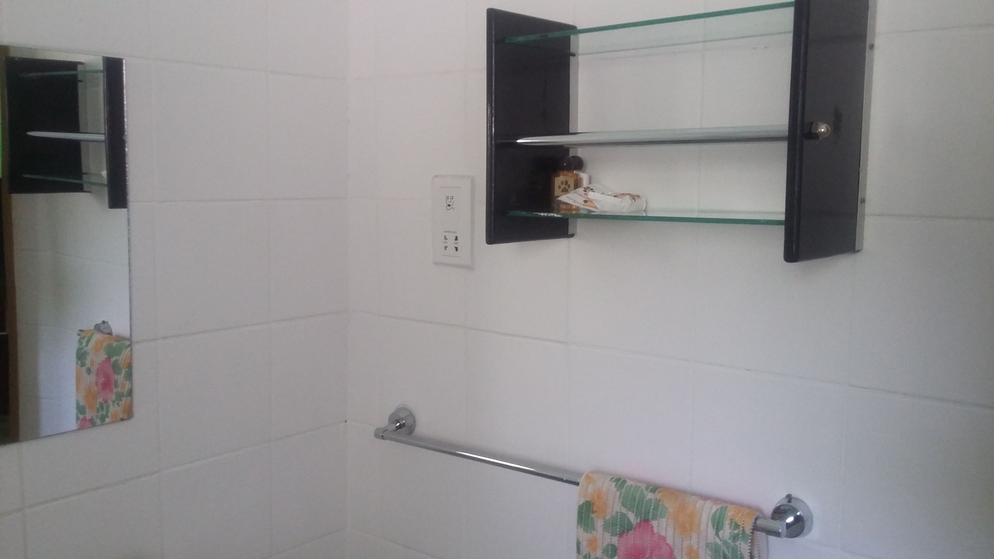 A bathroom with white tiled walls, a glass shelf with toiletries, a towel rack with a floral towel, and a mirror with a black frame.