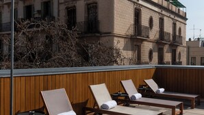 Una terraza en la azotea con tumbonas, un muro de madera y un edificio con balcones al fondo.